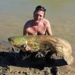 Glad fiskare stoltserar med en enorm mal fisk vid strandkanten.